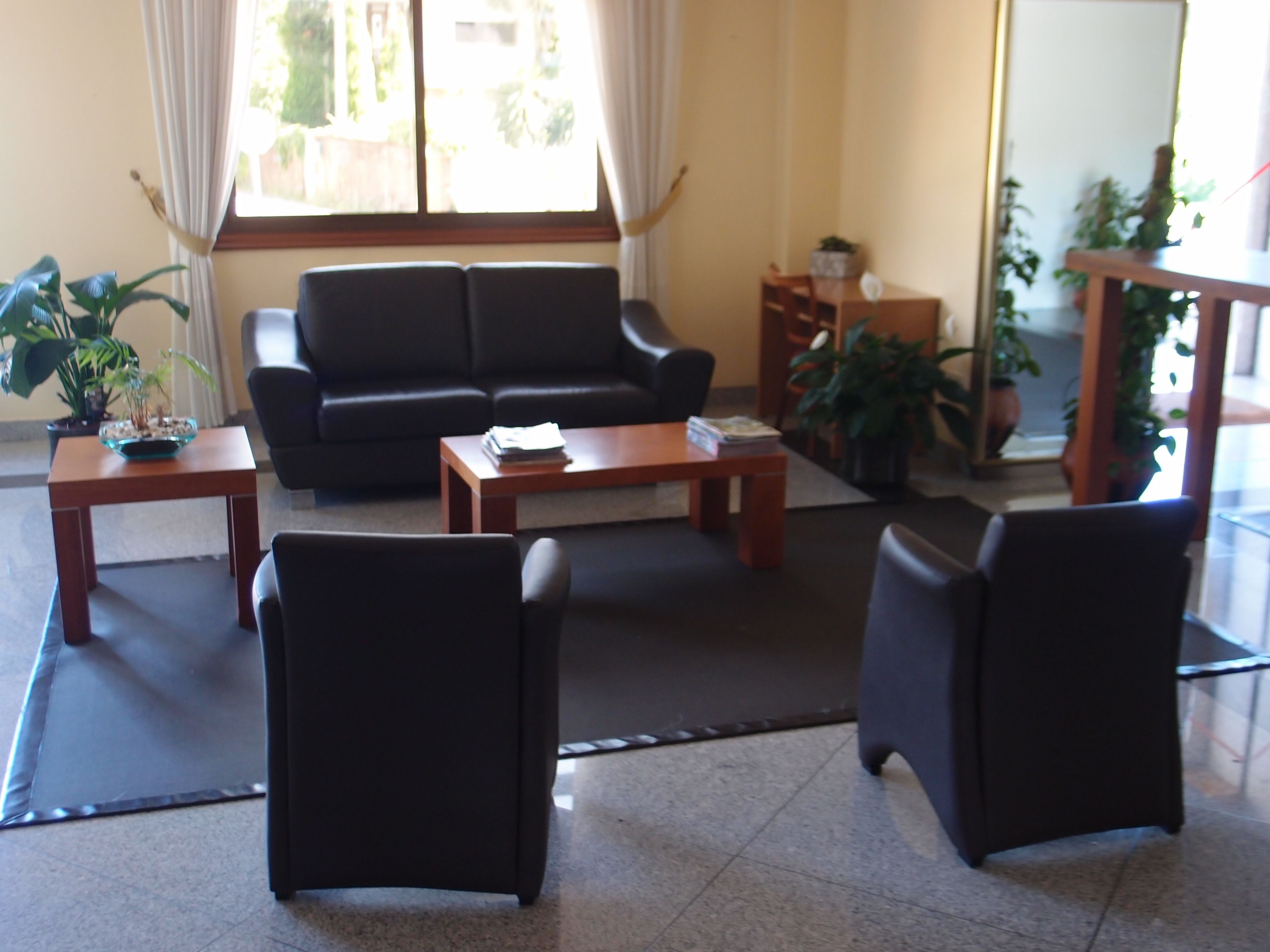Uma sala de estar com sofá, duas poltronas, uma mesa de centro e uma mesinha lateral. Há plantas em vasos e uma janela com cortinas.