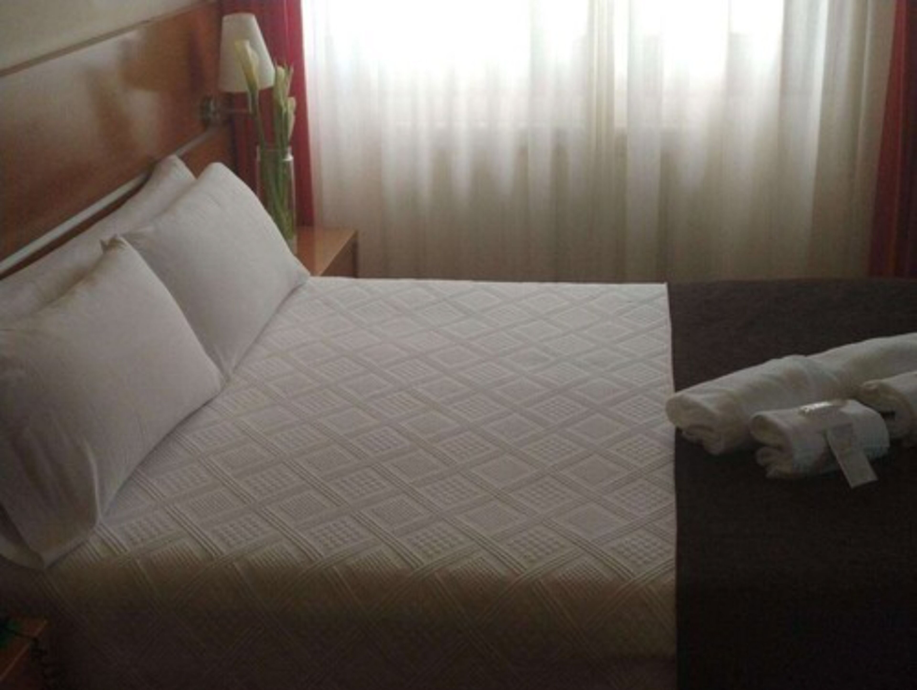 Ein Hotelzimmer mit einem großen Bett, einem Nachttisch mit Lampe und einem Fenster mit transparenten Vorhängen.