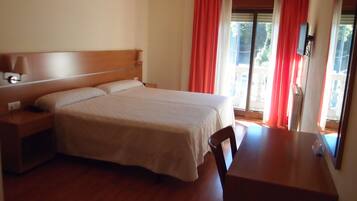 Habitación de hotel con una cama grande, una mesita de noche de madera, un escritorio y una silla. Hay una ventana con cortinas rojas y vistas al exterior.