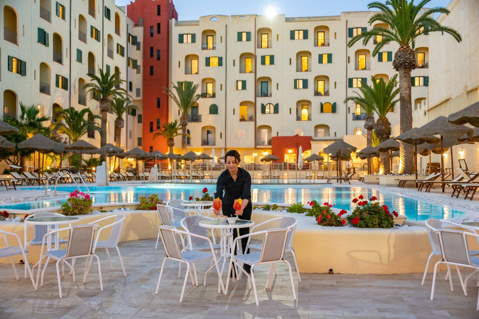 Zona piscina di un hotel con posti a sedere all'aperto e un edificio con numerosi balconi sullo sfondo.