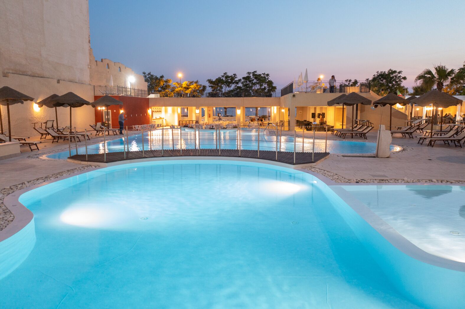 A swimming pool with lounge chairs and umbrellas, surrounded by a building with multiple levels and a balcony.