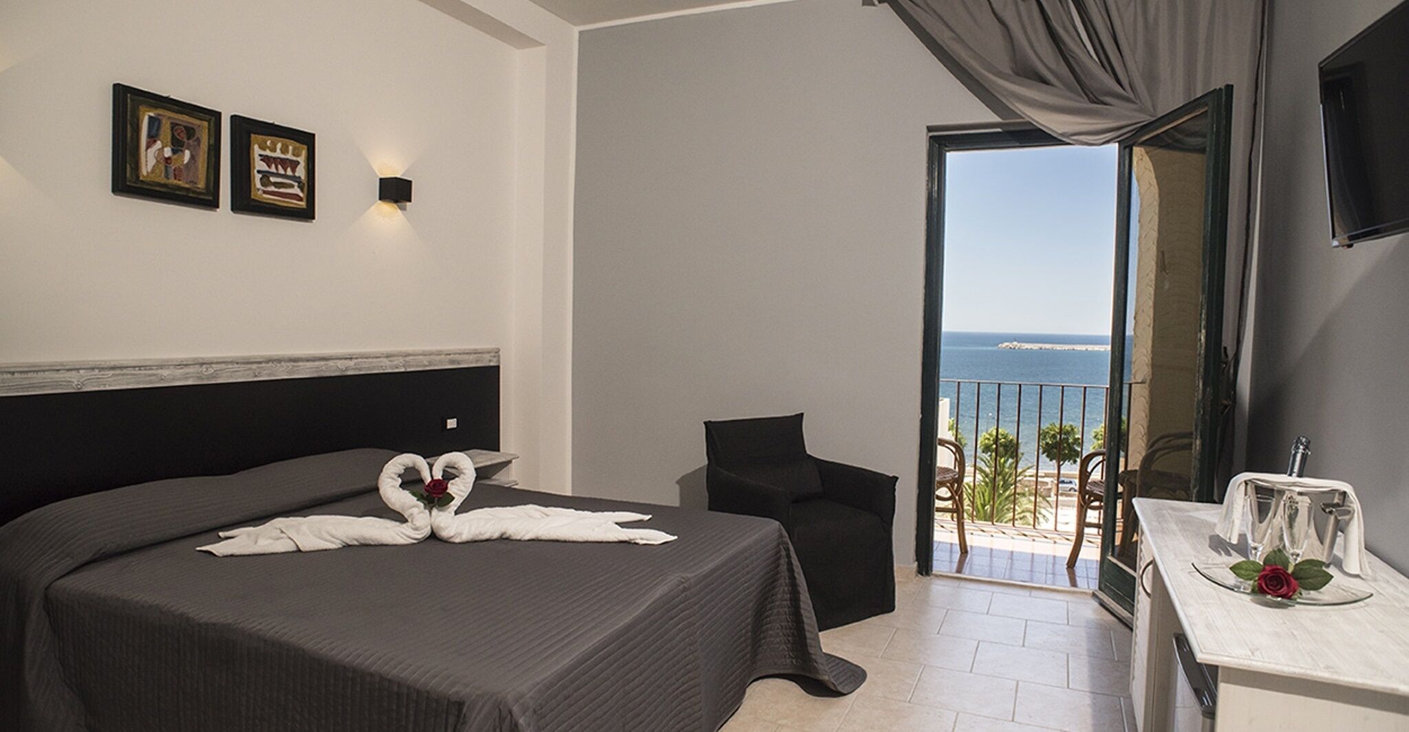 Een hotelkamer met een bed, een stoel, een balkon met uitzicht op zee en een tafel met glazen en een roos.
