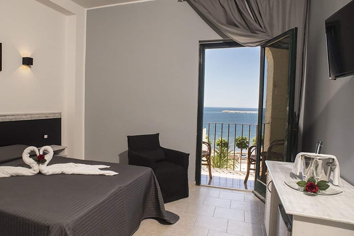 Habitación de hotel con cama, una silla, balcón con vista al mar y una mesa con copas y una rosa.