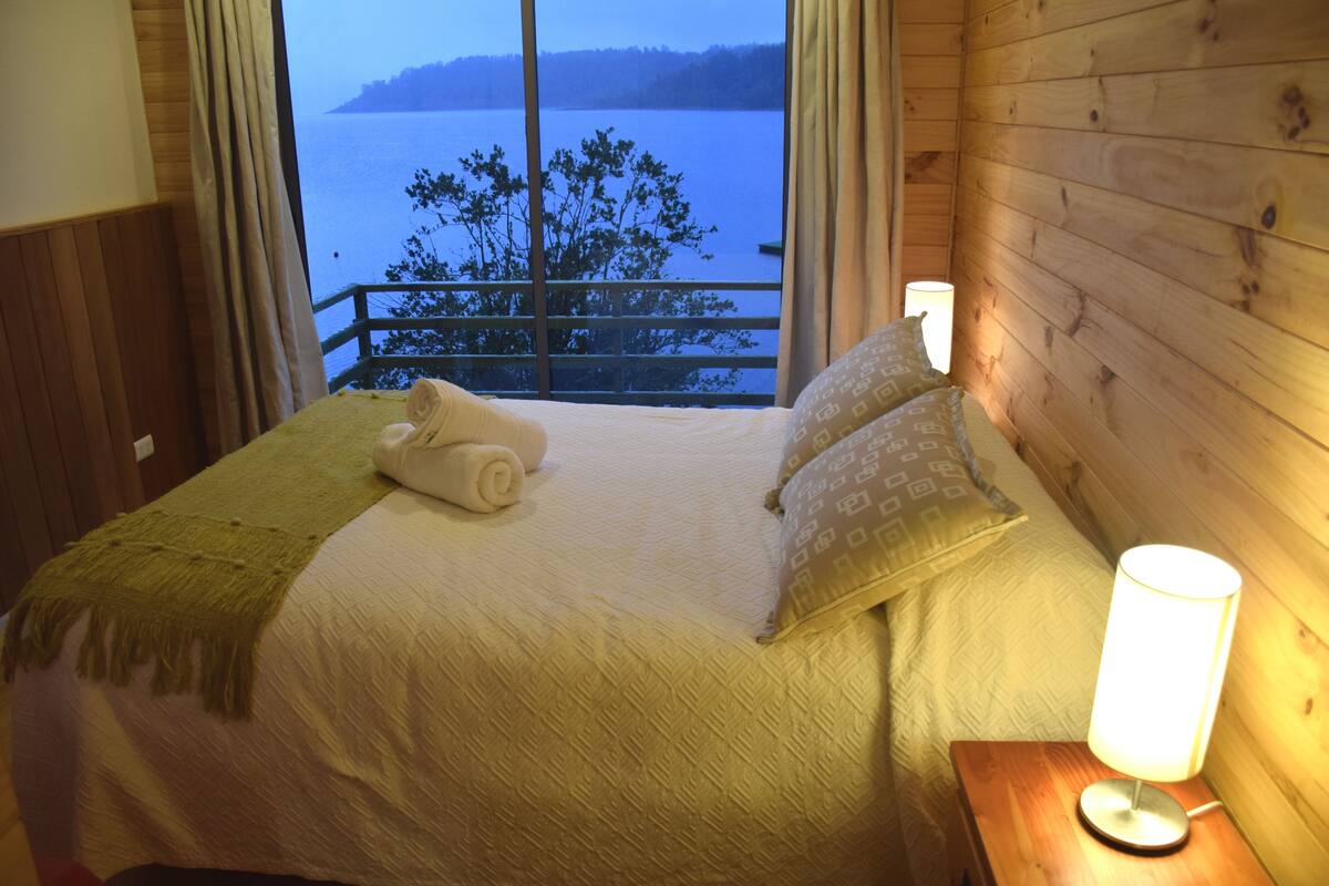 A bedroom with a bed, a nightstand with a lamp, a window with a view of a lake and trees, and a wooden wall.