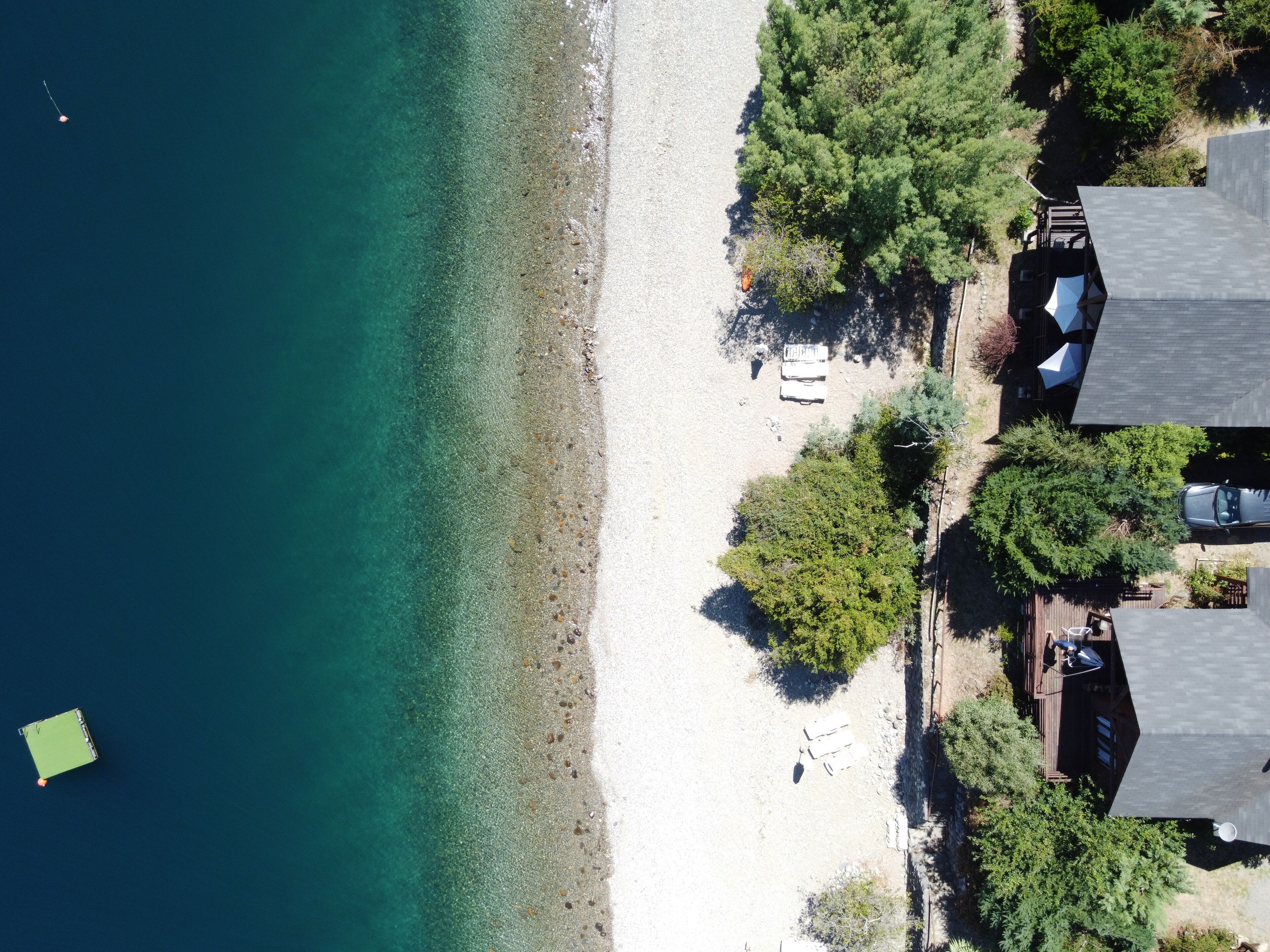 An aerial view of a beach with clear turquoise water, a sandy shore, and several buildings with visible outdoor seating areas.