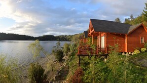 En træhytte med terrasse, beliggende ved en sø med udsigt til en skovklædt bakke.