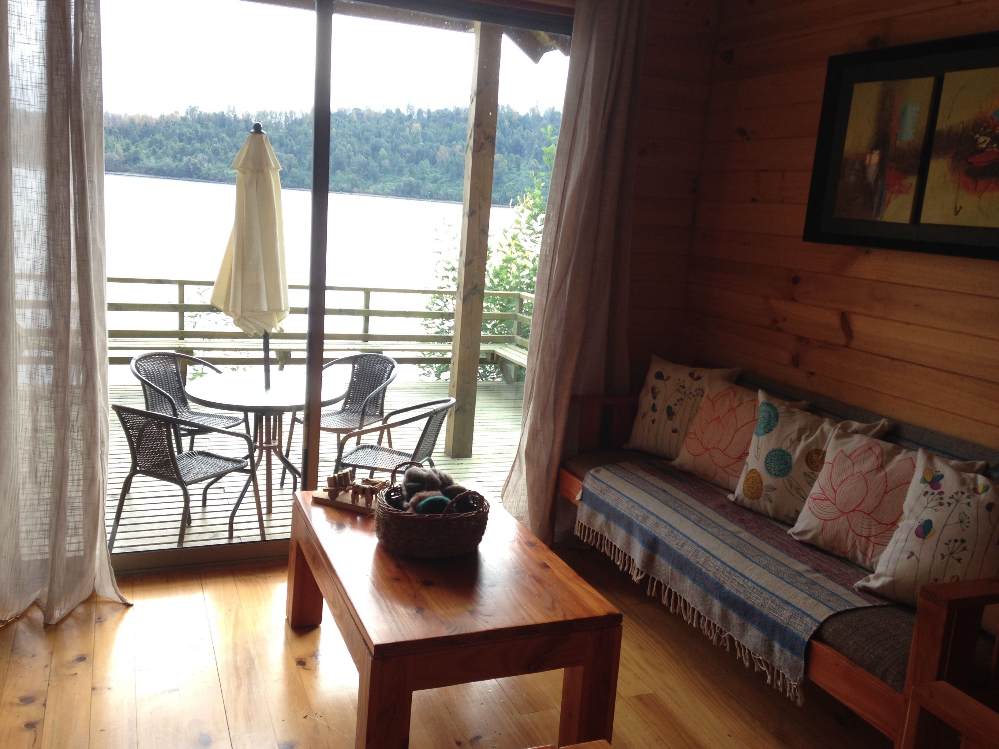 Eine Holzhütte mit Holztisch und -stühlen, einer Couch mit gemusterten Kissen und Blick auf einen See durch große Fenster.