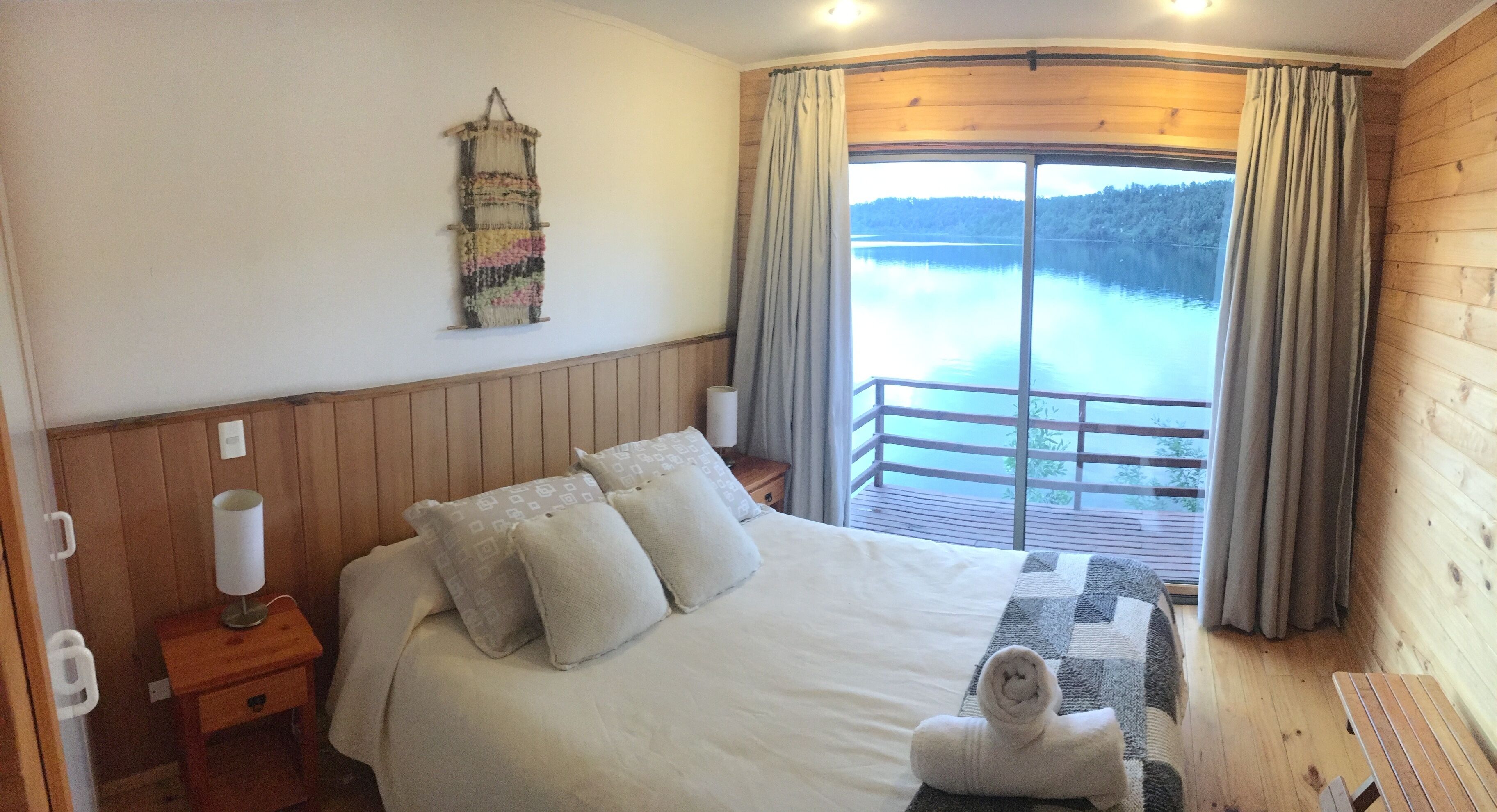 Ein Schlafzimmer mit einem großen Bett, einem Nachttisch mit Lampe, einer Wanddekoration und Blick auf einen See vom Balkon aus.