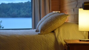 En seng med mønstret sengetøj, et natbord med lampe, og et vindue med udsigt over sø og skov.