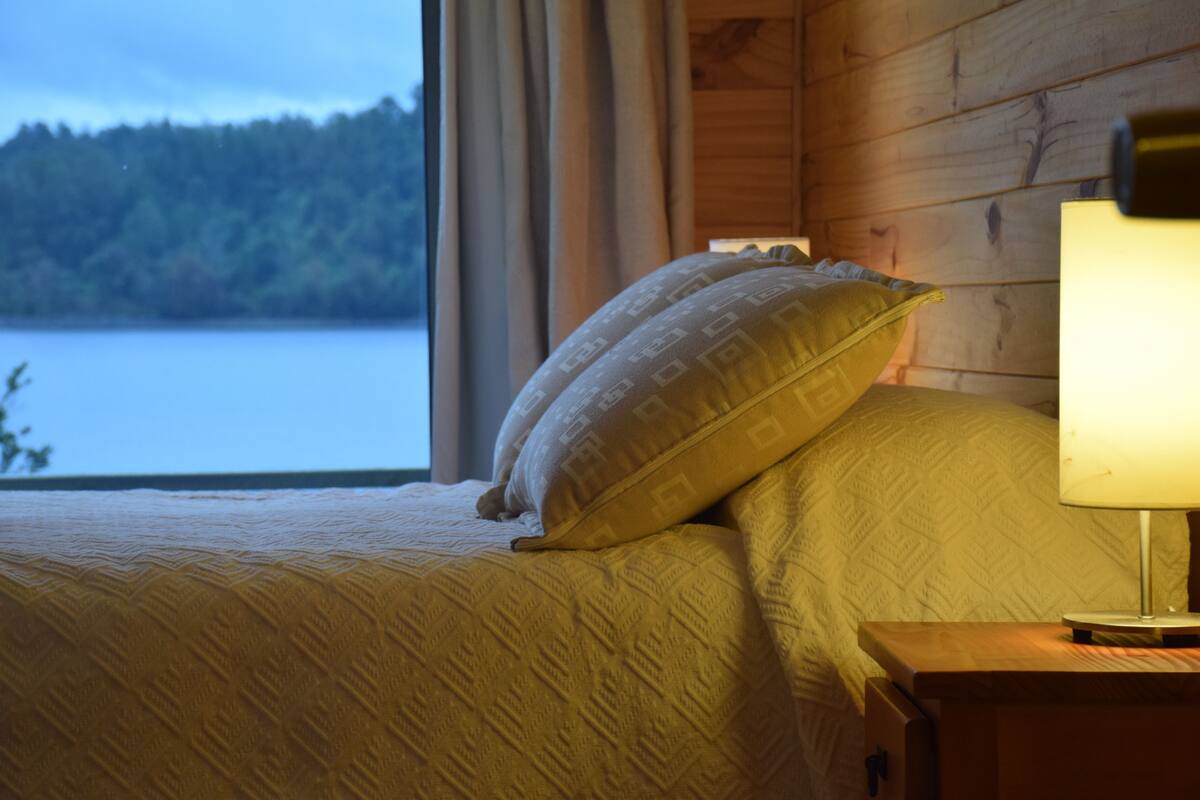 A bed with patterned bedding, a nightstand with a lamp, and a window with a view of a lake and forest.