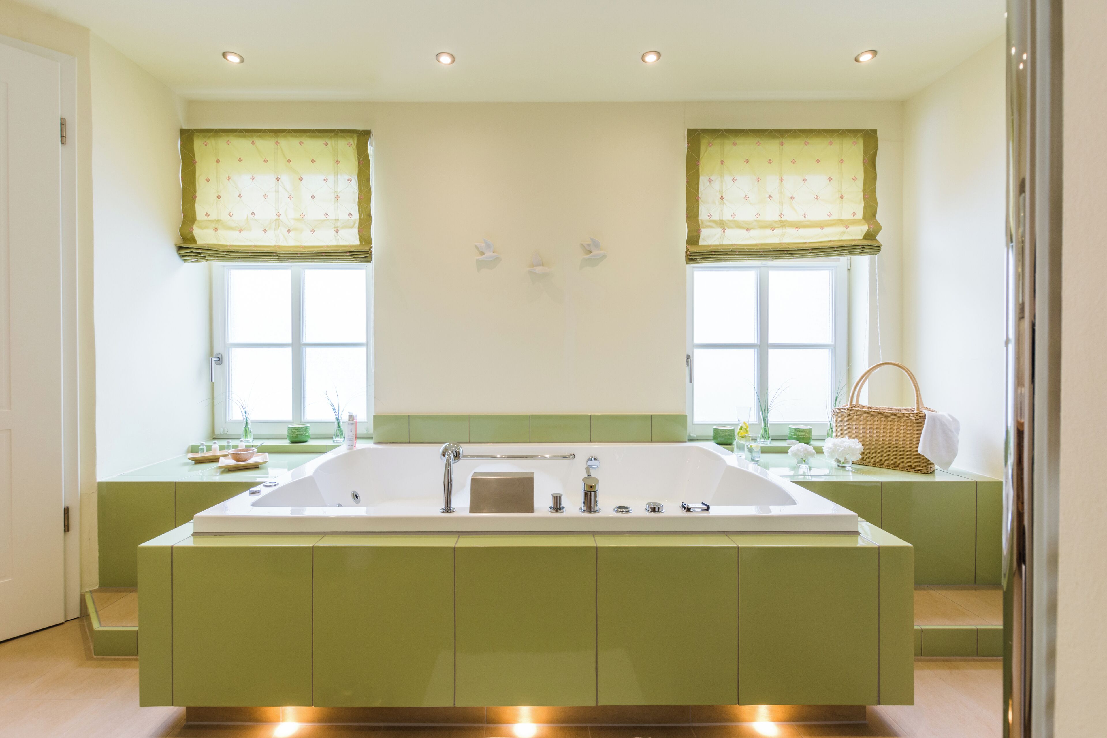 Un baño moderno con una bañera grande, muebles de color verde y dos ventanas con cortinas verdes.