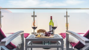 Een tafel met een fles wijn, twee glazen, een bord met kaas en druiven, en een mand met brood op een balkon met uitzicht op zee.