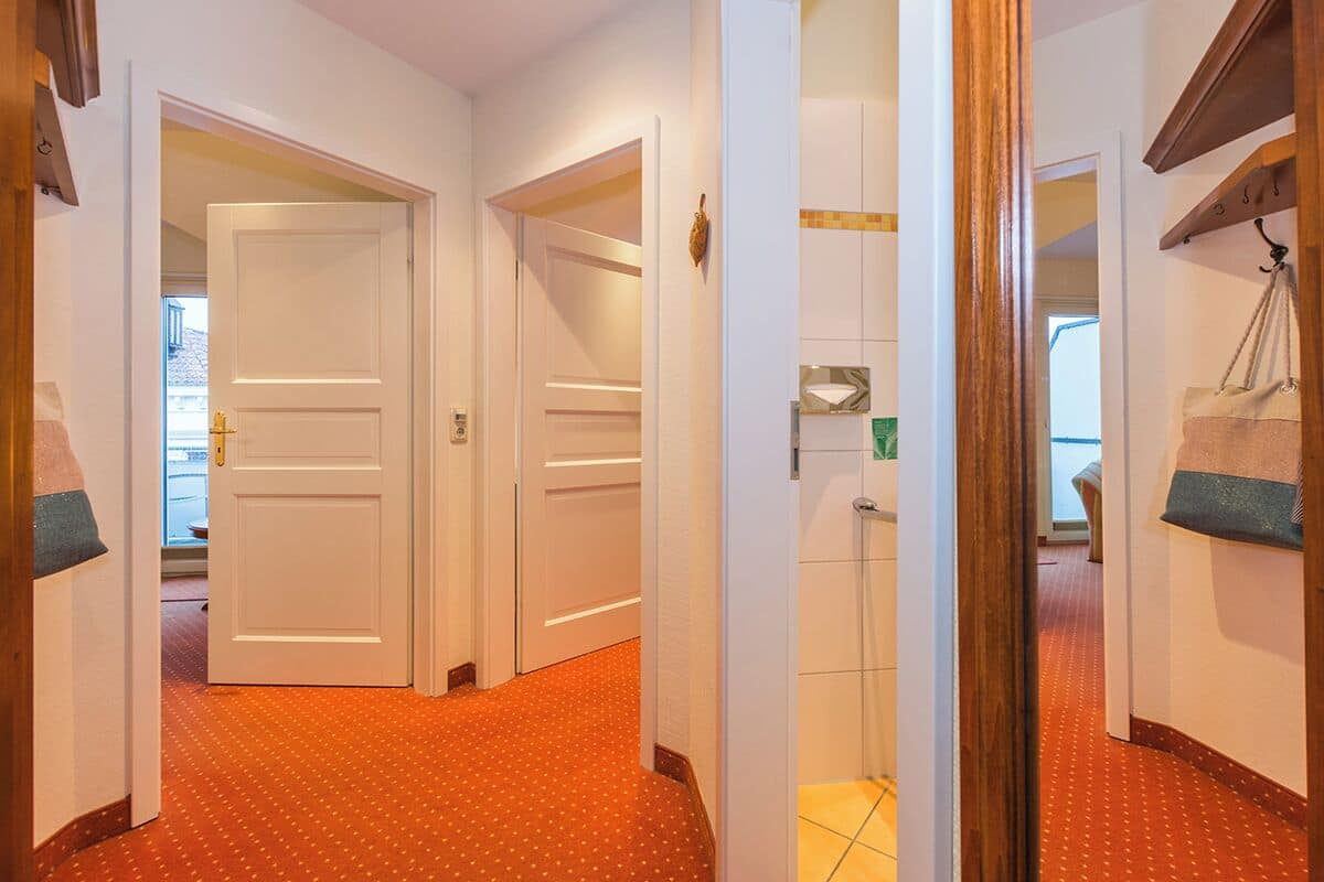 Un couloir d’hôtel avec un tapis rouge à motifs, des portes blanches et un banc en bois sur lequel sont posés des sacs.