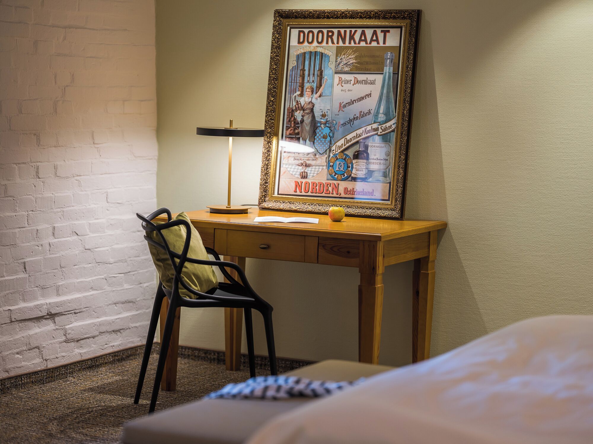 A hotel room with a wooden desk, a chair, a framed picture on the wall, and a bed in the foreground.