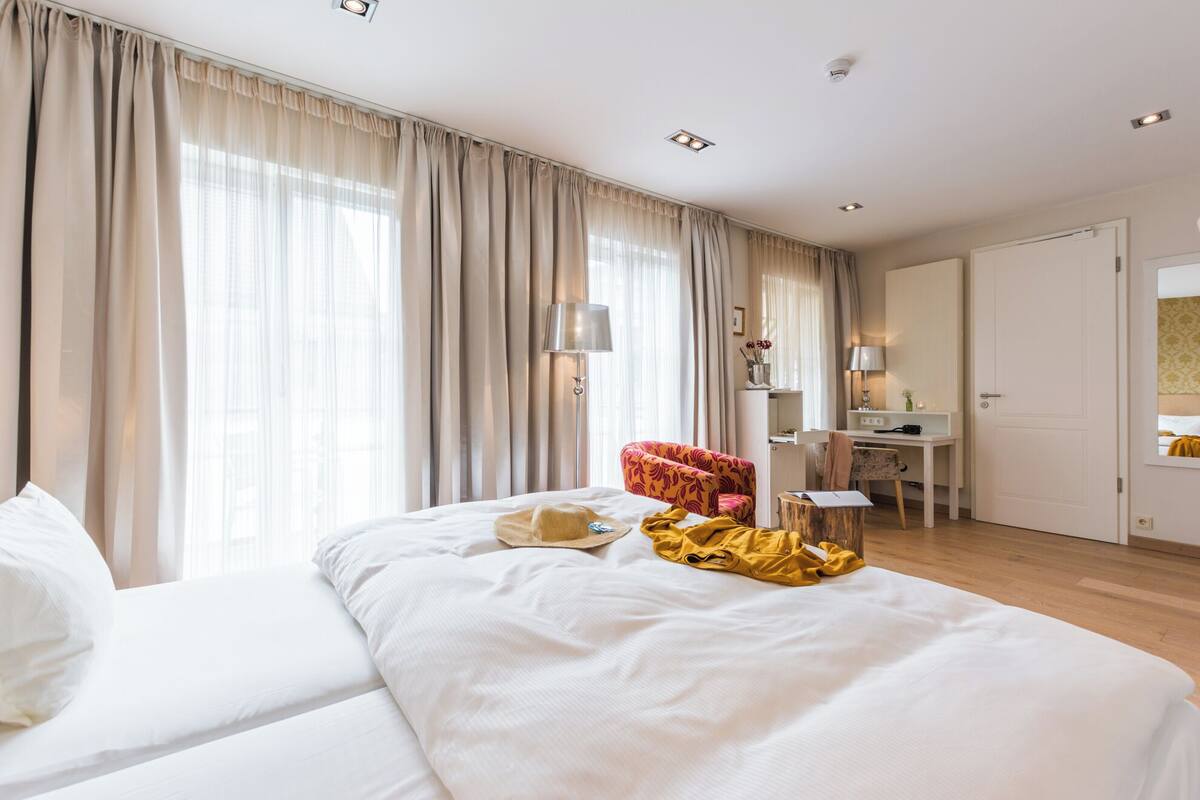 Une chambre d’hôtel avec un grand lit, un bureau et une chaise. Il y a des rideaux aux fenêtres et une lampe sur le bureau.