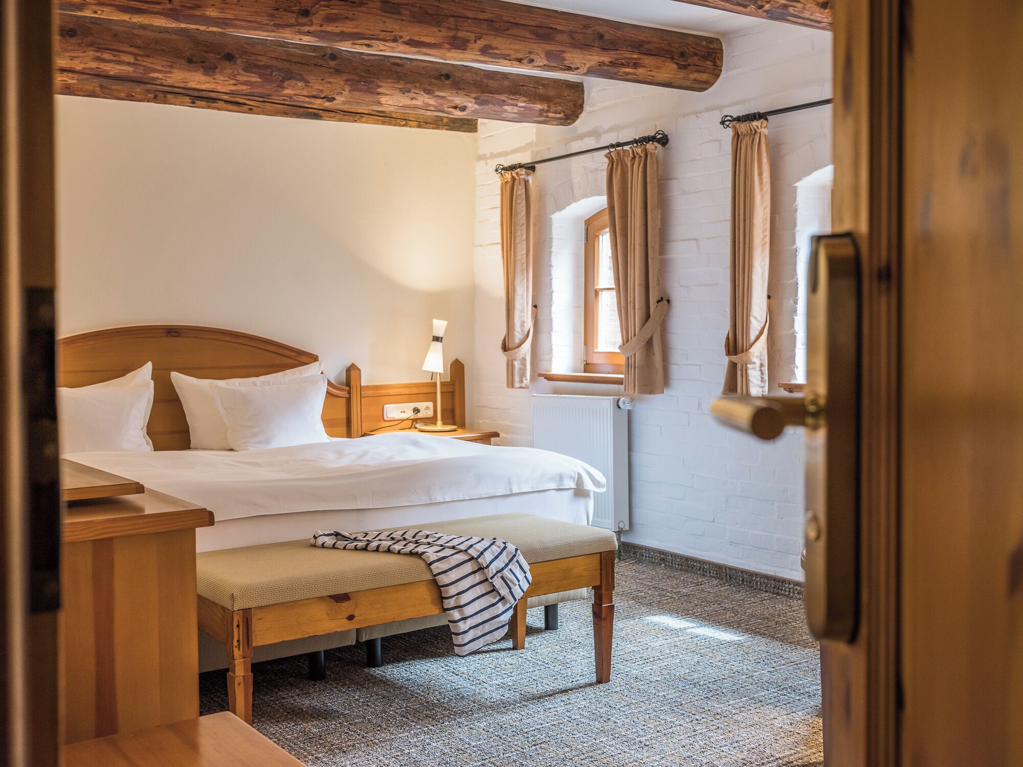 A bedroom with a wooden bed, a bedside table with a lamp, a bench, and a window with curtains.