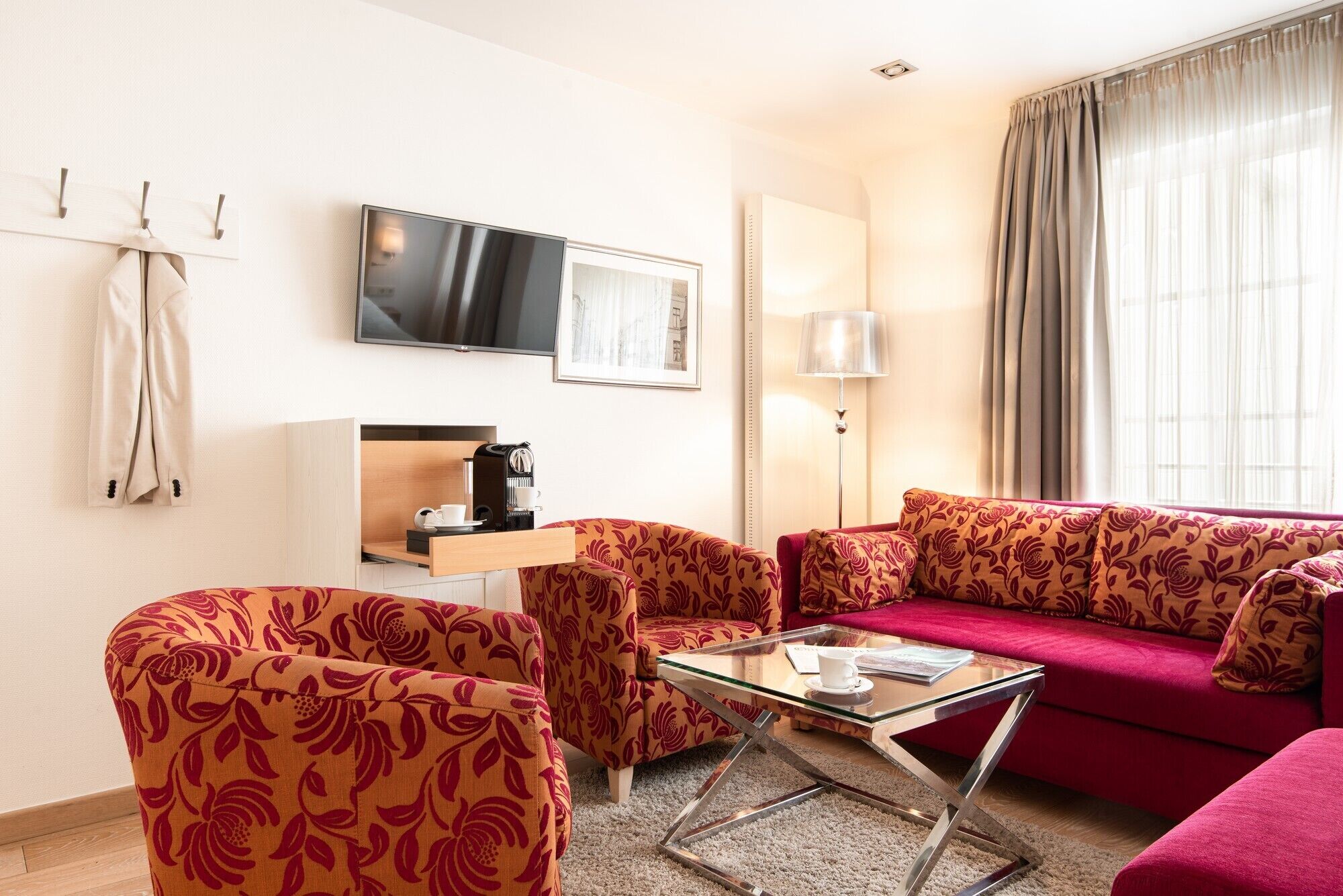 A modern living room with a sofa, armchairs, a coffee table, a flat-screen TV, a coffee machine, and a lamp.