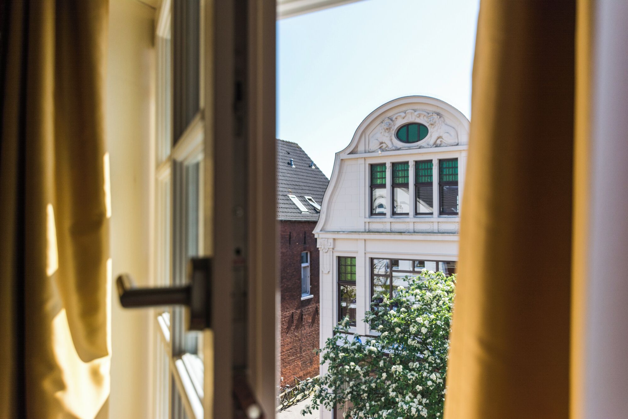 An interior view of a room with a window looking out to a white building with green shutters and a white door.