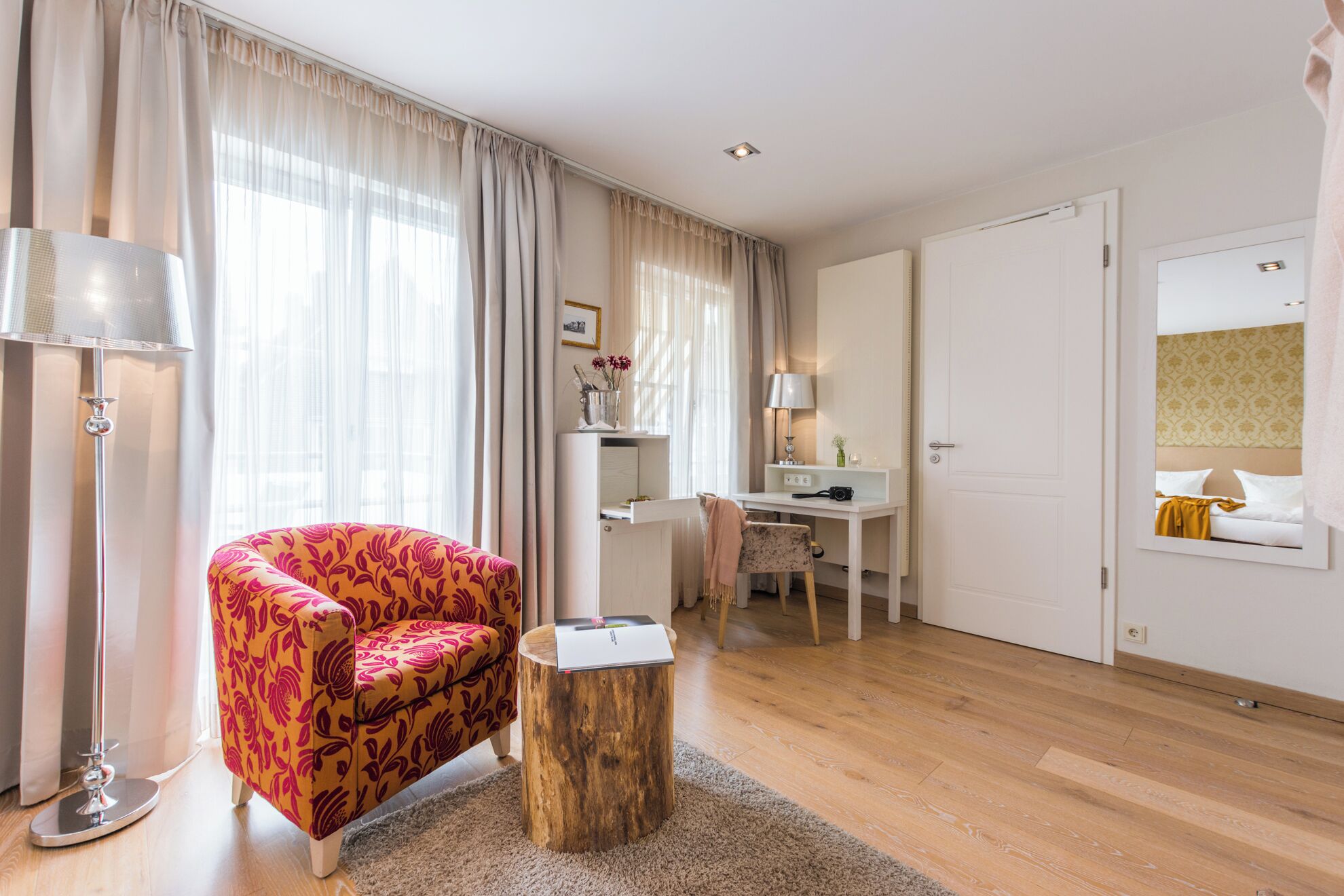 Una habitación de hotel moderna con suelo de madera, un sillón tapizado con estampado floral rojo, una mesita de madera pequeña, un escritorio con lámpara y una cama con cabecero.