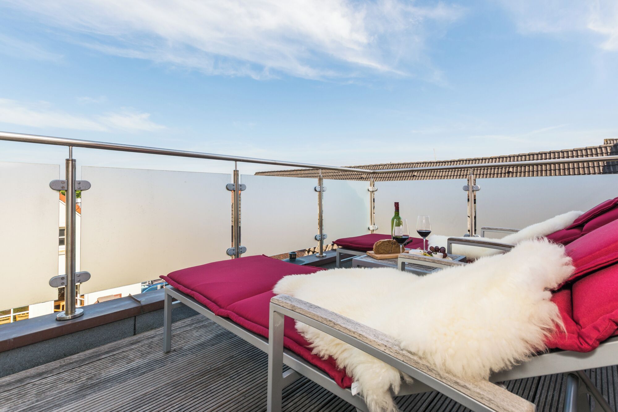 Une terrasse sur le toit avec un banc recouvert de fourrure blanche, des coussins rouges et une table sur laquelle se trouvent une bouteille et des verres.