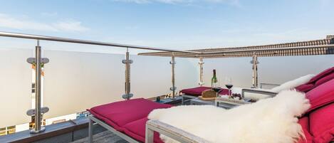 Une terrasse sur le toit avec un banc recouvert de fourrure blanche, des coussins rouges et une table sur laquelle se trouvent une bouteille et des verres.