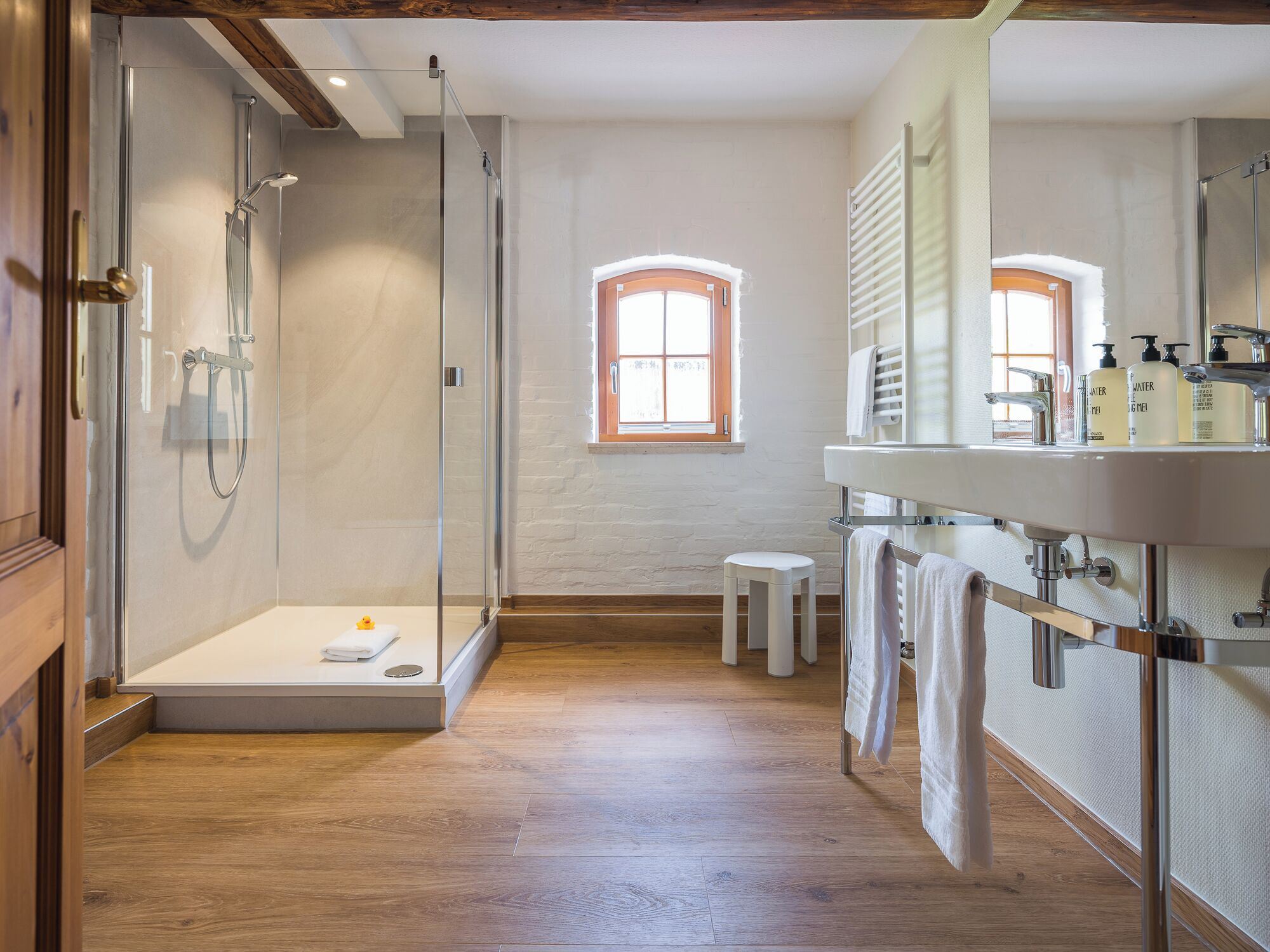A modern bathroom with a glass-enclosed shower, a wooden floor, a small stool, and a vanity with a mirror and toiletries.