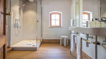 Um banheiro moderno com chuveiro em cabine de vidro, piso de madeira, um banquinho pequeno e um espelho com produtos de higiene pessoal.