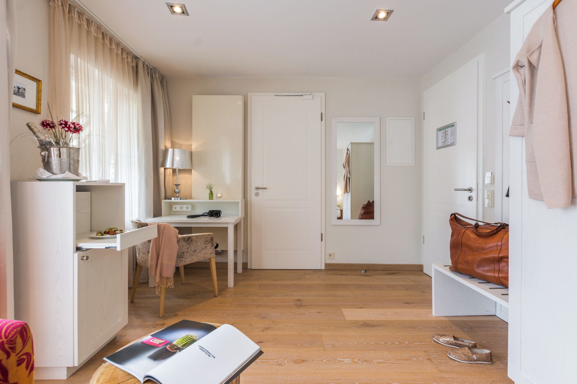 Habitación de hotel compacta con suelo de madera, un escritorio con lámpara, una silla, una mesita con un jarrón de flores, un espejo y una puerta que da a otra habitación.