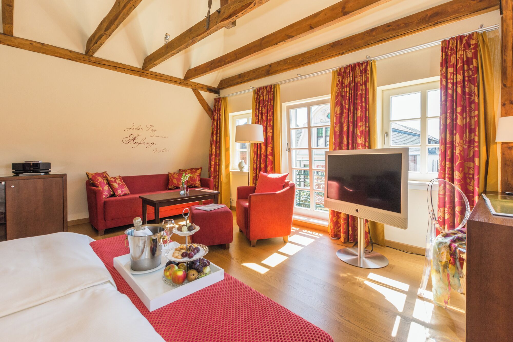 A cozy hotel room with a wooden ceiling, a sofa, a TV, and a dining area with a fruit display.