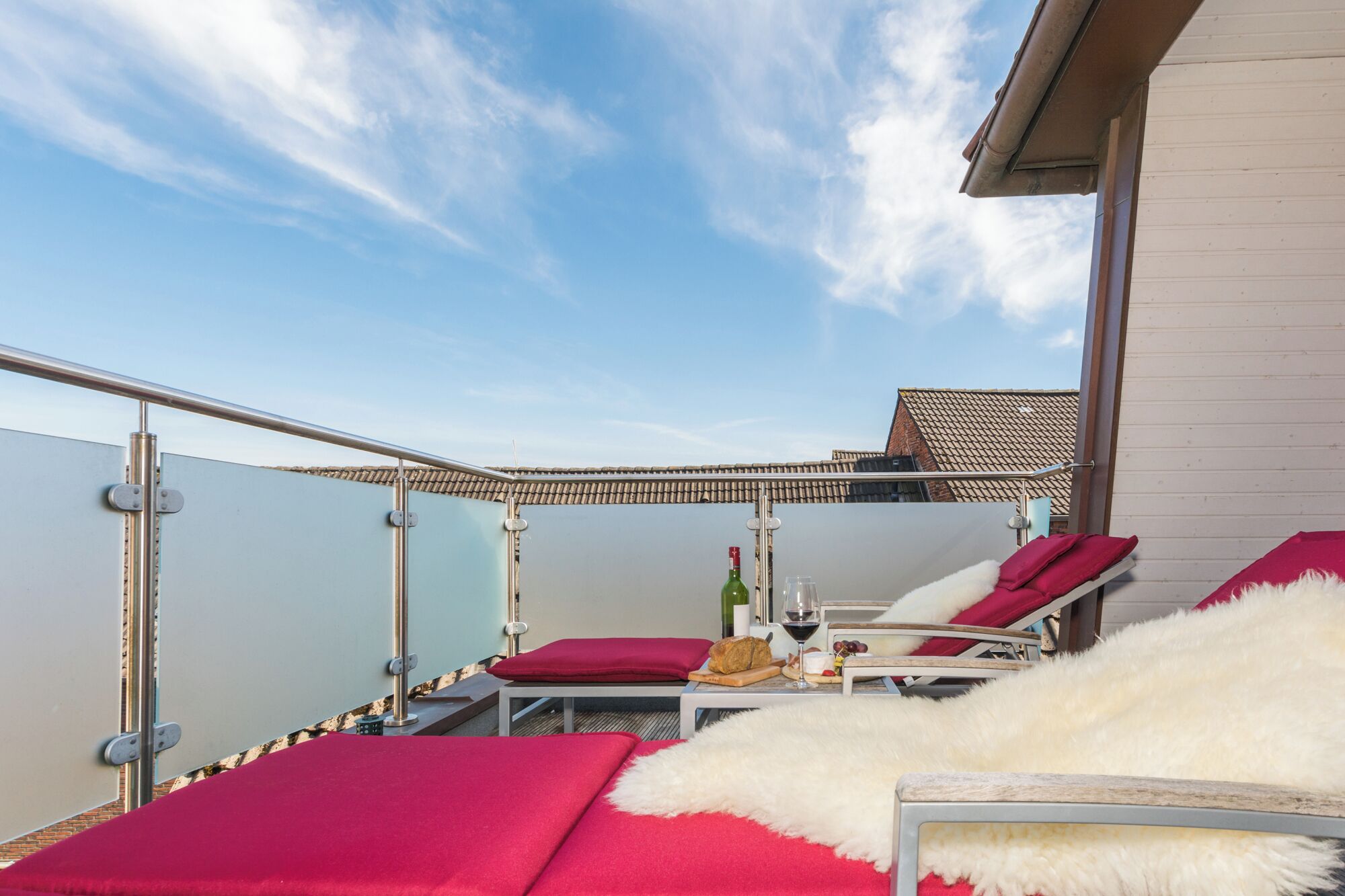 A balcony with red cushioned lounge chairs, a small table with a bottle and glasses, and a view of the sea.