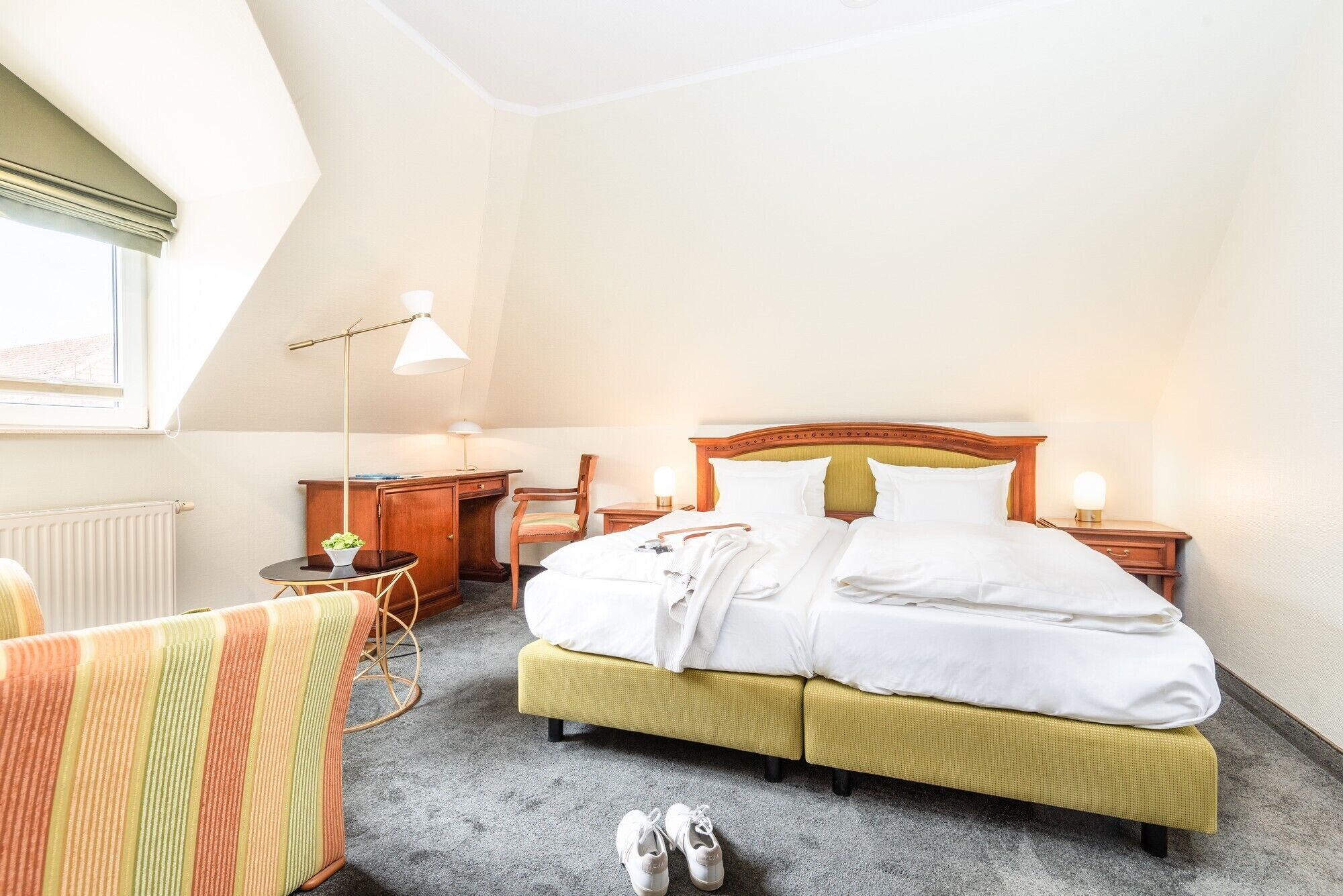 A hotel room with a bed, a desk, a chair, a lamp, and a radiator.