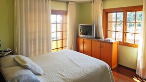 Un dormitorio con cama, cortinas, televisor sobre un mueble de madera y ventanas con persianas.