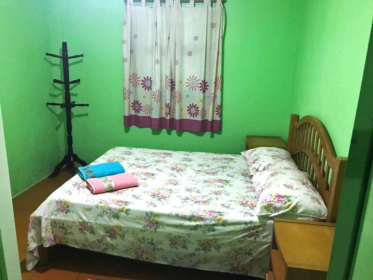 Een kleine kamer met een bed, een houten hoofdbord, een nachtkastje, een raam met gordijn en een kapstok.