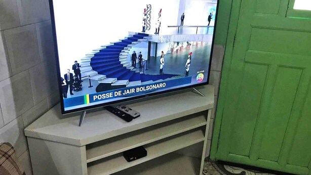 Een televisie op een witte standaard toont een nieuwsuitzending, met een trap en mensen.
