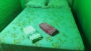 Een kleine kamer met een eenpersoonsbed, een bedovertrek met bloemenprint, een netjes gevouwen witte en roze handdoek en een raam met kantgordijnen.