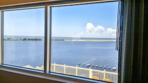 A view through a window with a railing, overlooking a large body of water and a distant shoreline with trees and buildings.