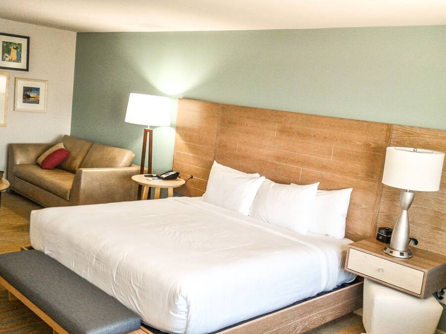 Habitación de hotel con una cama grande, una banca, una mesita de noche con lámpara, una pequeña mesa con cesta, un sofá con almohada roja y cuadros enmarcados en la pared.