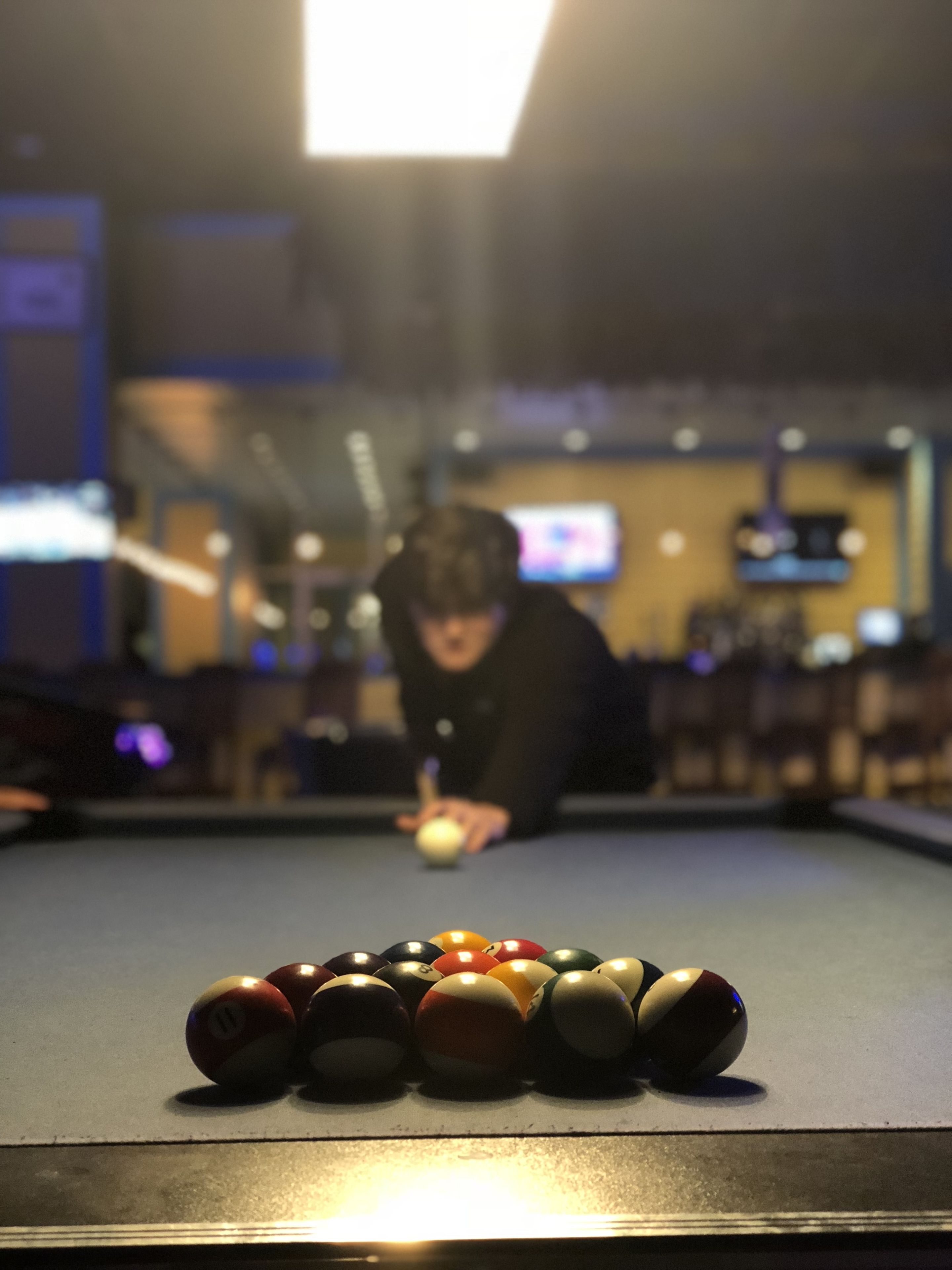Une table de billard avec les billes disposées en forme de triangle, une personne qui vise la bille blanche, et une pièce faiblement éclairée avec un téléviseur en arrière-plan.