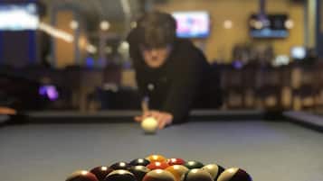 Una mesa de billar con bolas dispuestas en forma de triángulo, una persona apuntando a la bola blanca, y una habitación con poca luz y un televisor al fondo.