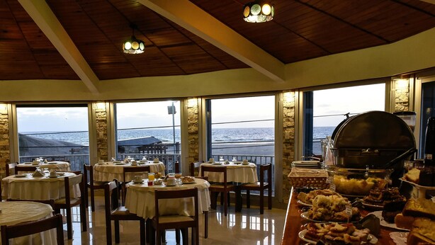A restaurant with round tables and chairs, a buffet with various dishes, and a view of the sea through large windows. - Hotel Altair (San Clemente)