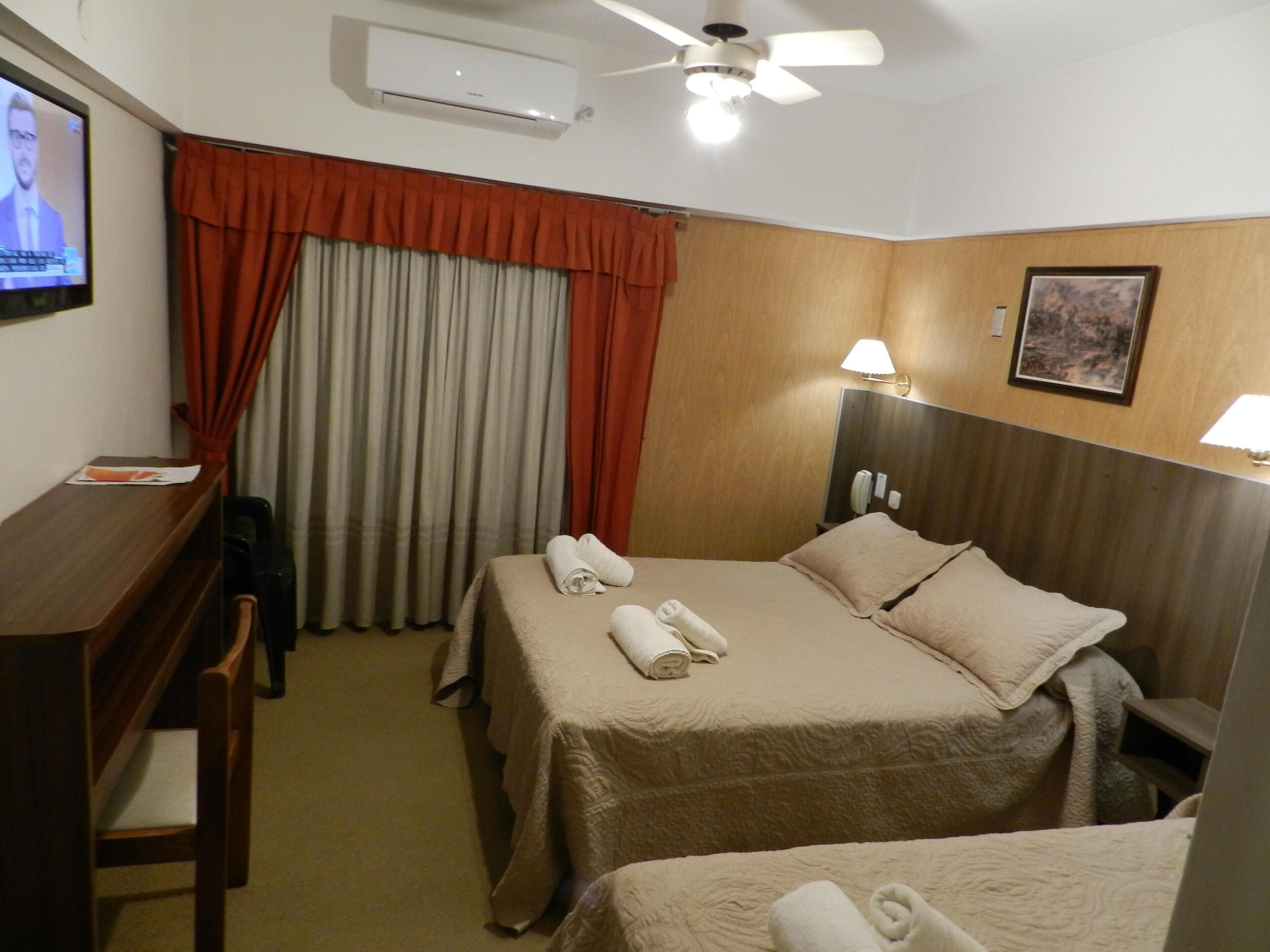 A hotel room with a bed, a desk, a chair, a TV, an air conditioner, a ceiling fan, and a painting on the wall.