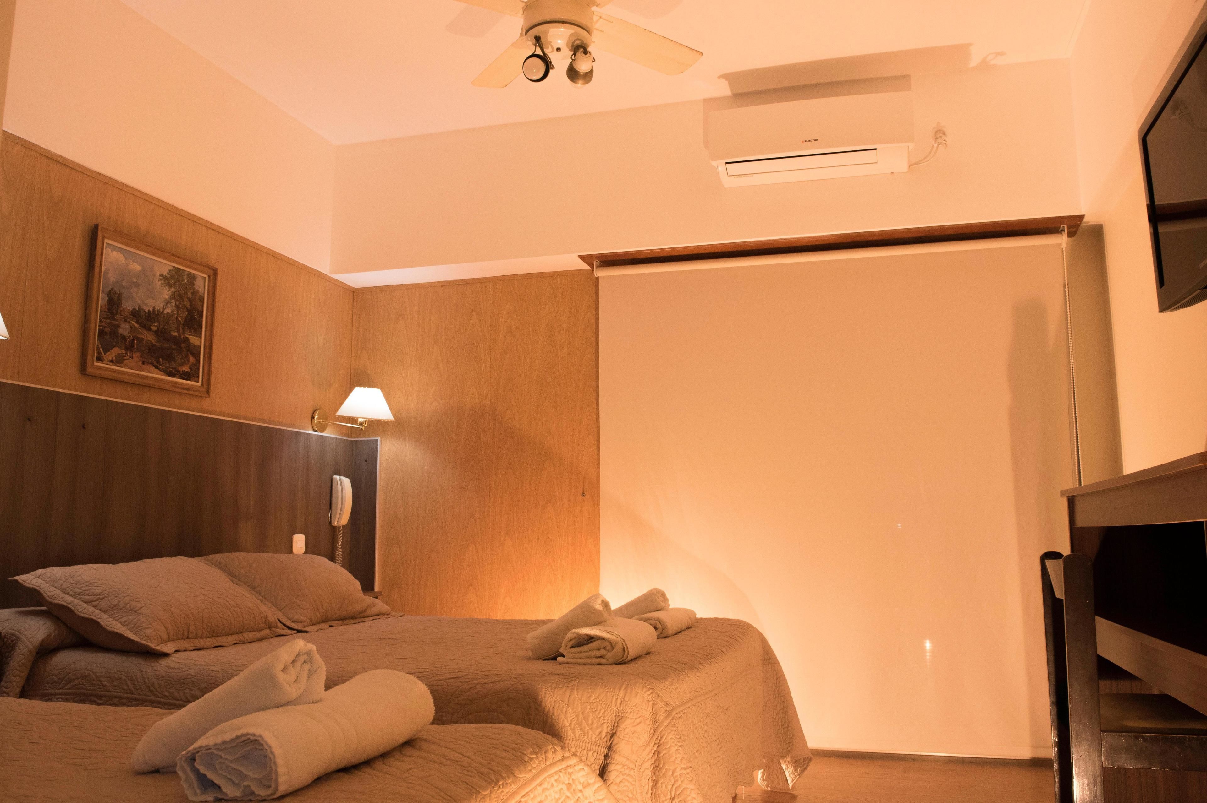 A hotel room with a bed, a ceiling fan, an air conditioning unit, a painting on the wall, and a telephone on the nightstand.