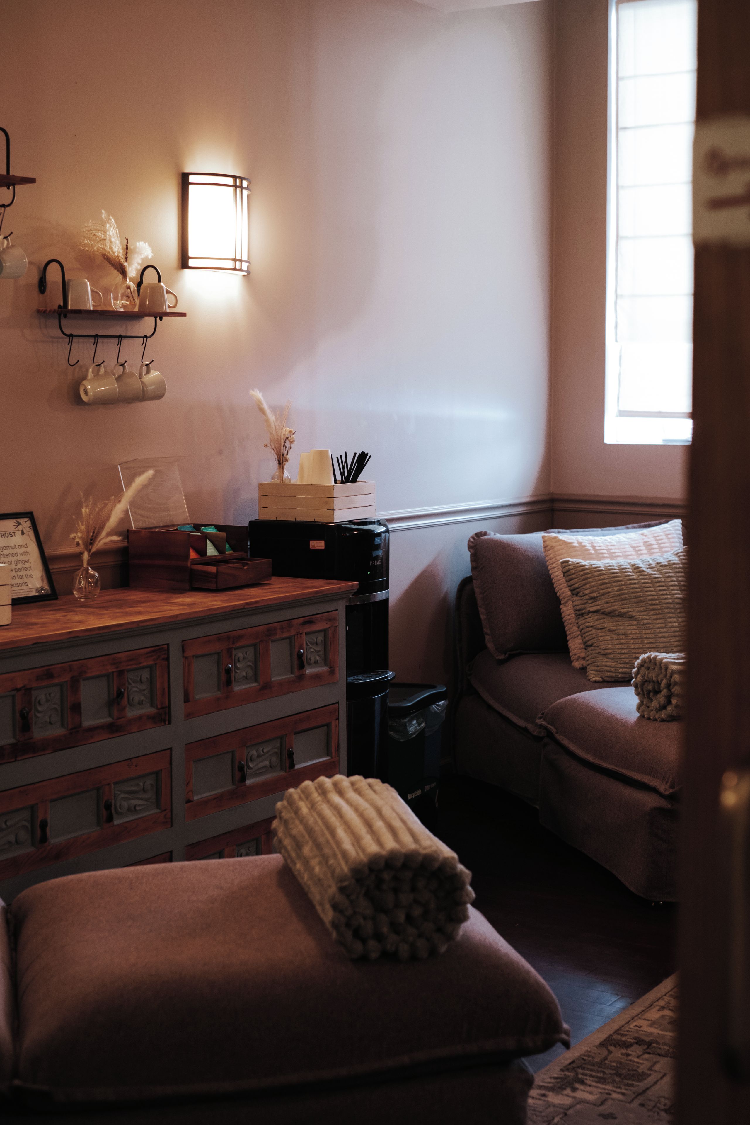 Una habitación acogedora con un aparador de madera, un banco acolchado, un estante en la pared con tazas y una ventana con persianas.