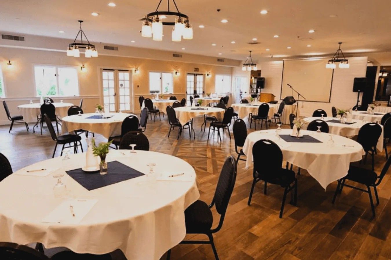 Une salle de banquet spacieuse, dotée de nombreuses tables rondes, de chaises et de luminaires au plafond.