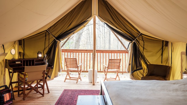 Een tentaccommodatie met een houten terras, twee stoelen, een bed en een bureau met een lamp.
