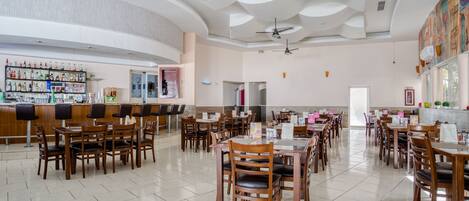 A spacious restaurant with multiple dining tables and chairs, a bar area with a variety of beverages, and ceiling fans.