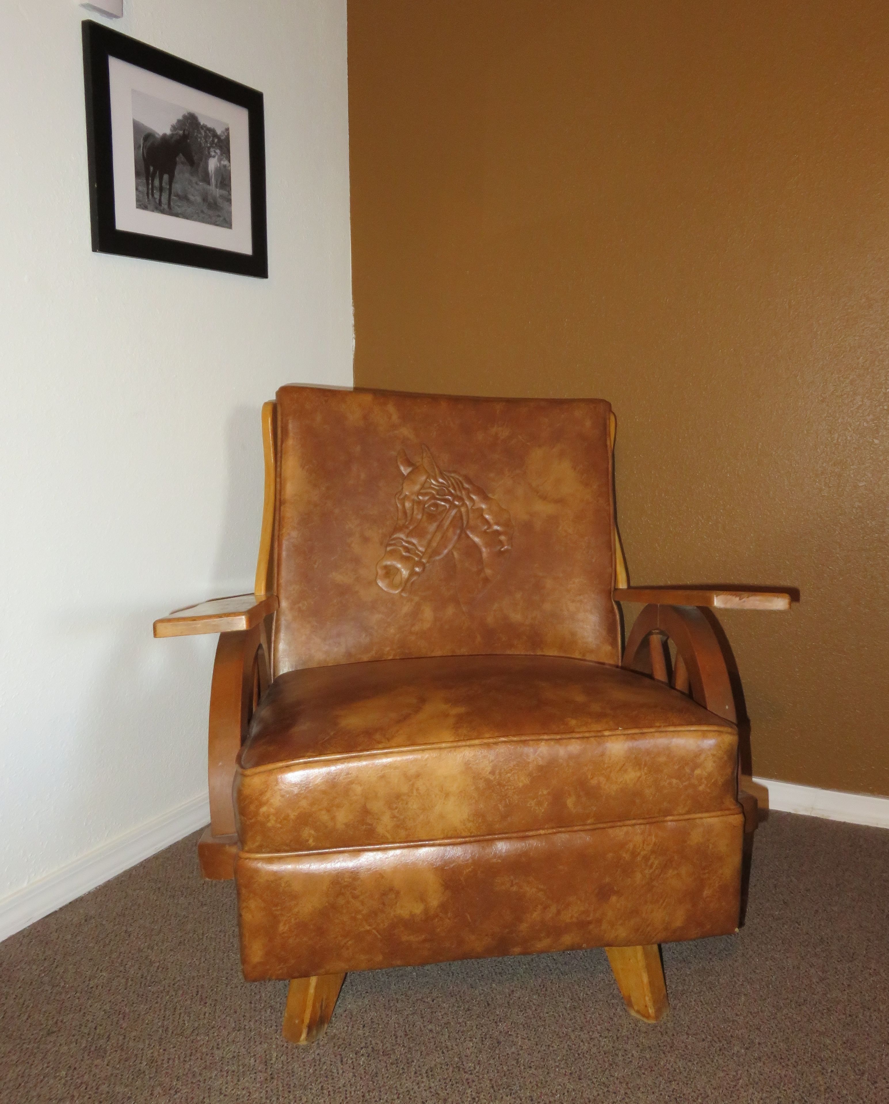 A brown leather armchair with a horse design on the backrest, positioned against a white wall with a framed picture above it.