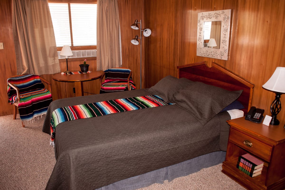 A hotel room with a bed, a desk, a chair, a lamp, and a bedside table with a phone and books.