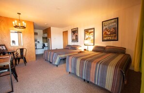 A hotel room with two beds, a dining area with chairs, a kitchen in the background, and a poster on the wall.