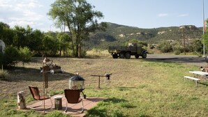 A rustic outdoor setting with a picnic table, chairs, and a bird feeder. A military vehicle is parked in the background.